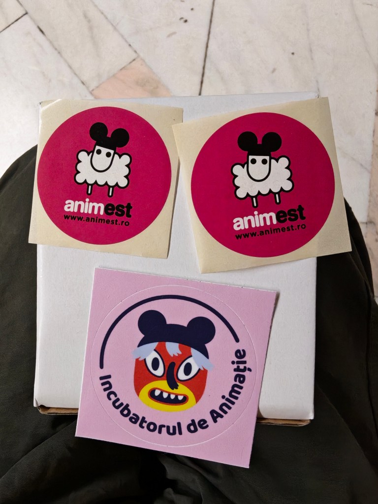 Două autocolante ale festivalului Anim'est, cu un personaj amuzant și logo-ul website-ului, și un autocolant denumit 'Incubatorul de Animație' cu un design creativ.